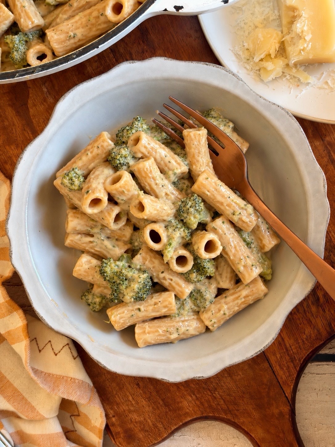 Rigatoni pasta with broccoli in a creamy sauce in a bowl with a fork, next to a block of parmesan cheese.