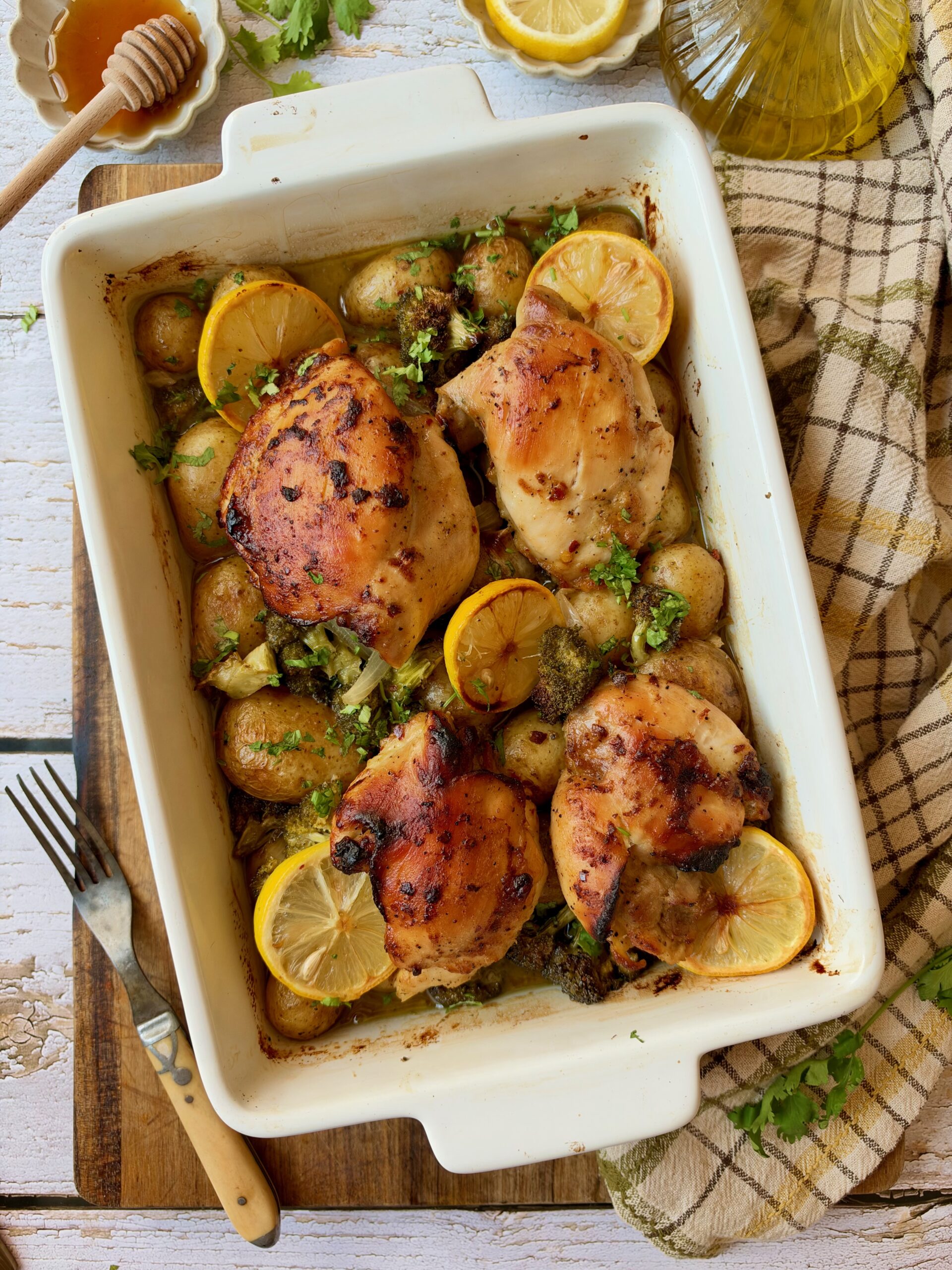 Honey mustard chicken thighs baked with potatoes, broccoli, and lemon slices in a white baking dish.