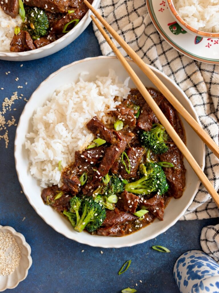 Crockpot Mongolian beef and broccoli served over white rice with chopsticks, topped with sesame seeds and green onions.