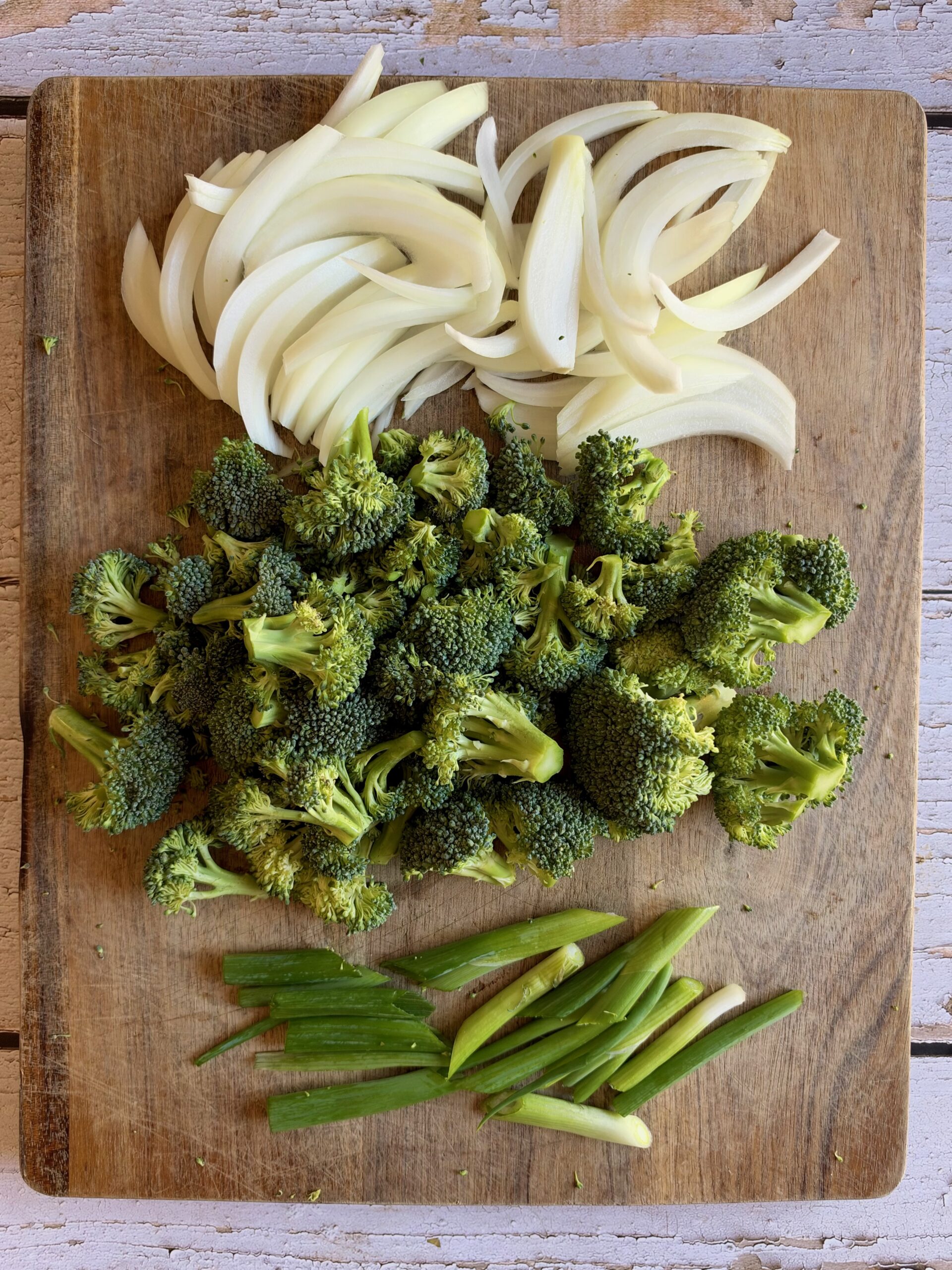 Sliced yellow onion, broccoli florets, and green onions on a wooden cutting board.