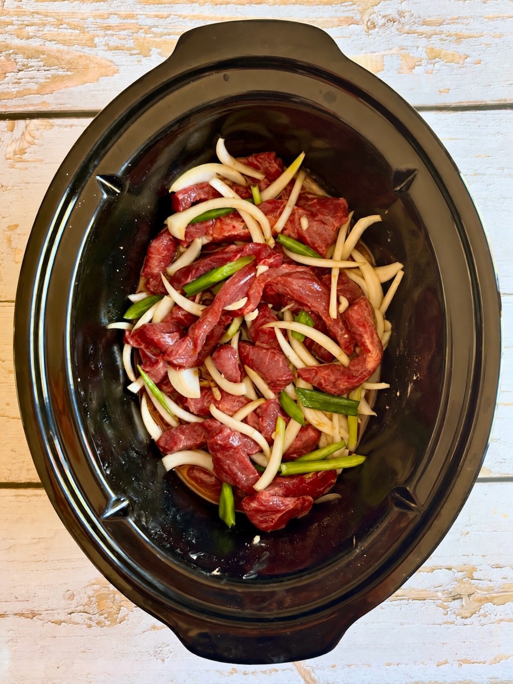 Cornstarch-coated beef with sliced onion and green onions in a slow cooker.