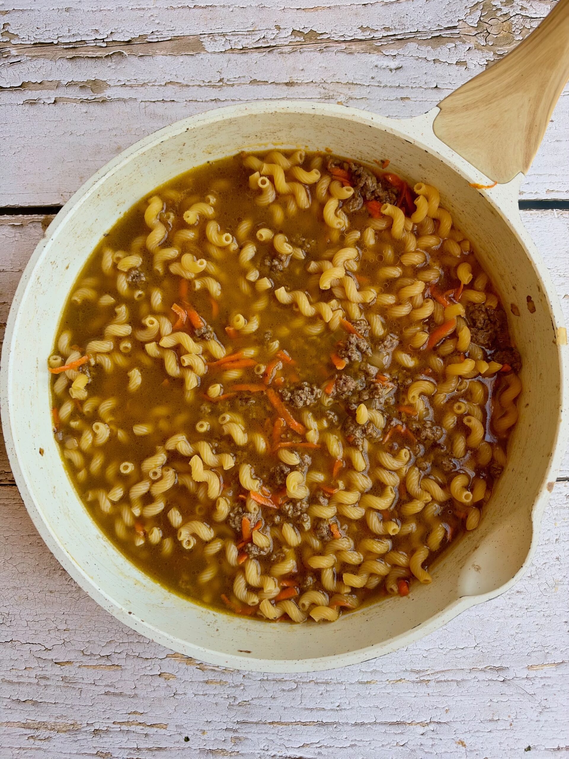 Uncooked pasta simmering in broth with ground beef and grated carrots in a white skillet.