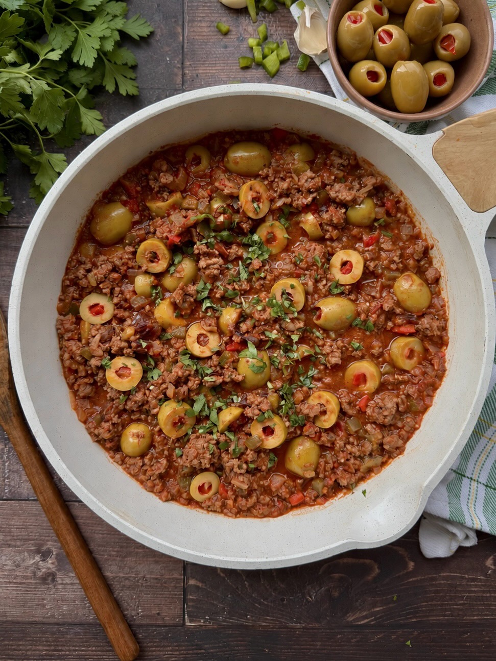Finished picadillo with sliced green olives and fresh parsley in a white skillet.