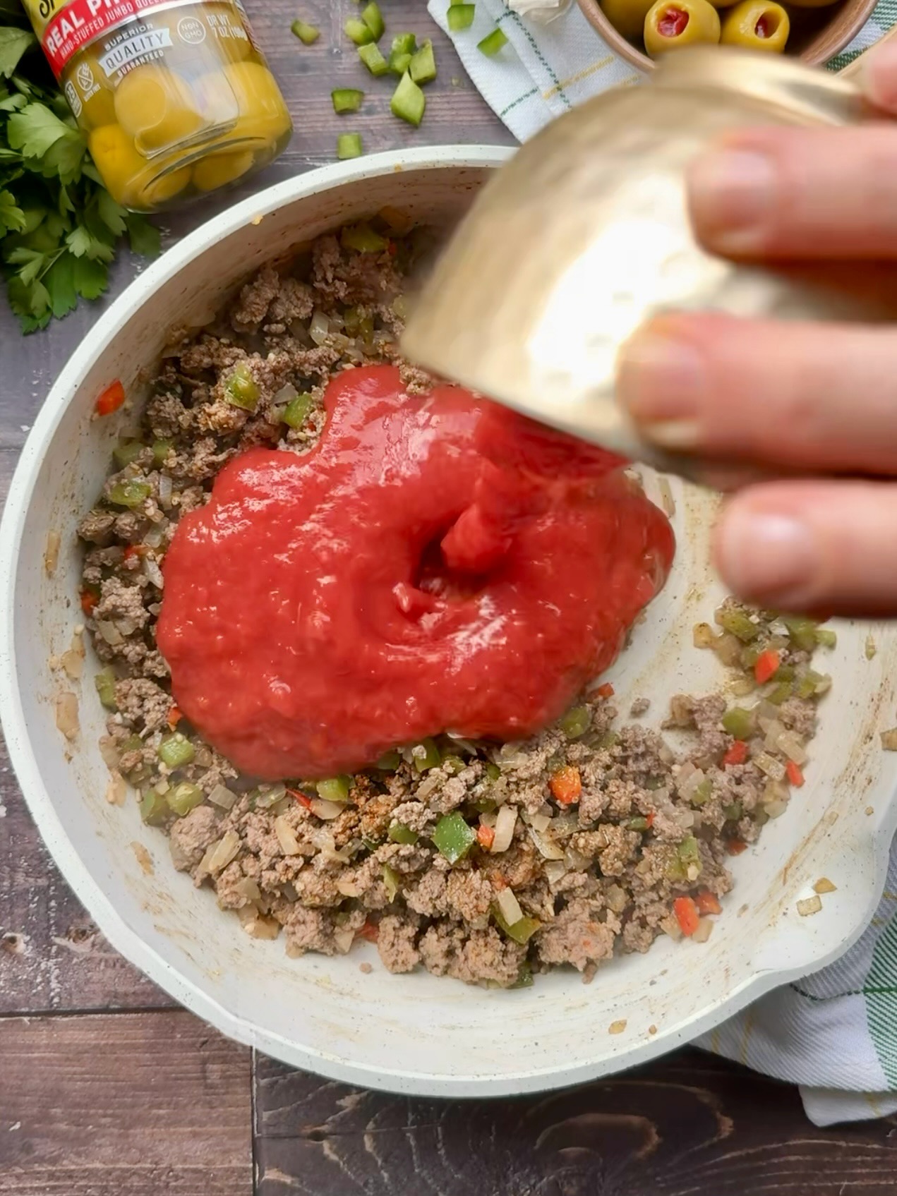 Tomato sauce being poured over browned ground beef in a white skillet.