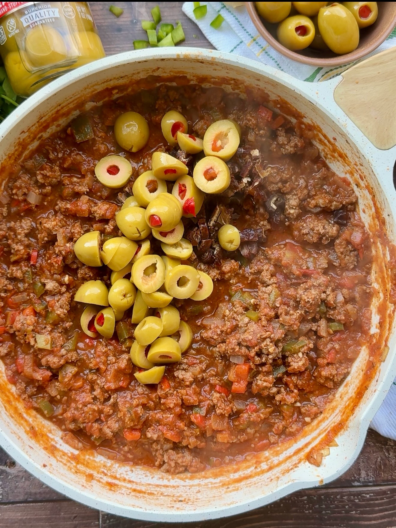 Sliced pimiento-stuffed olives added to picadillo in a white skillet.