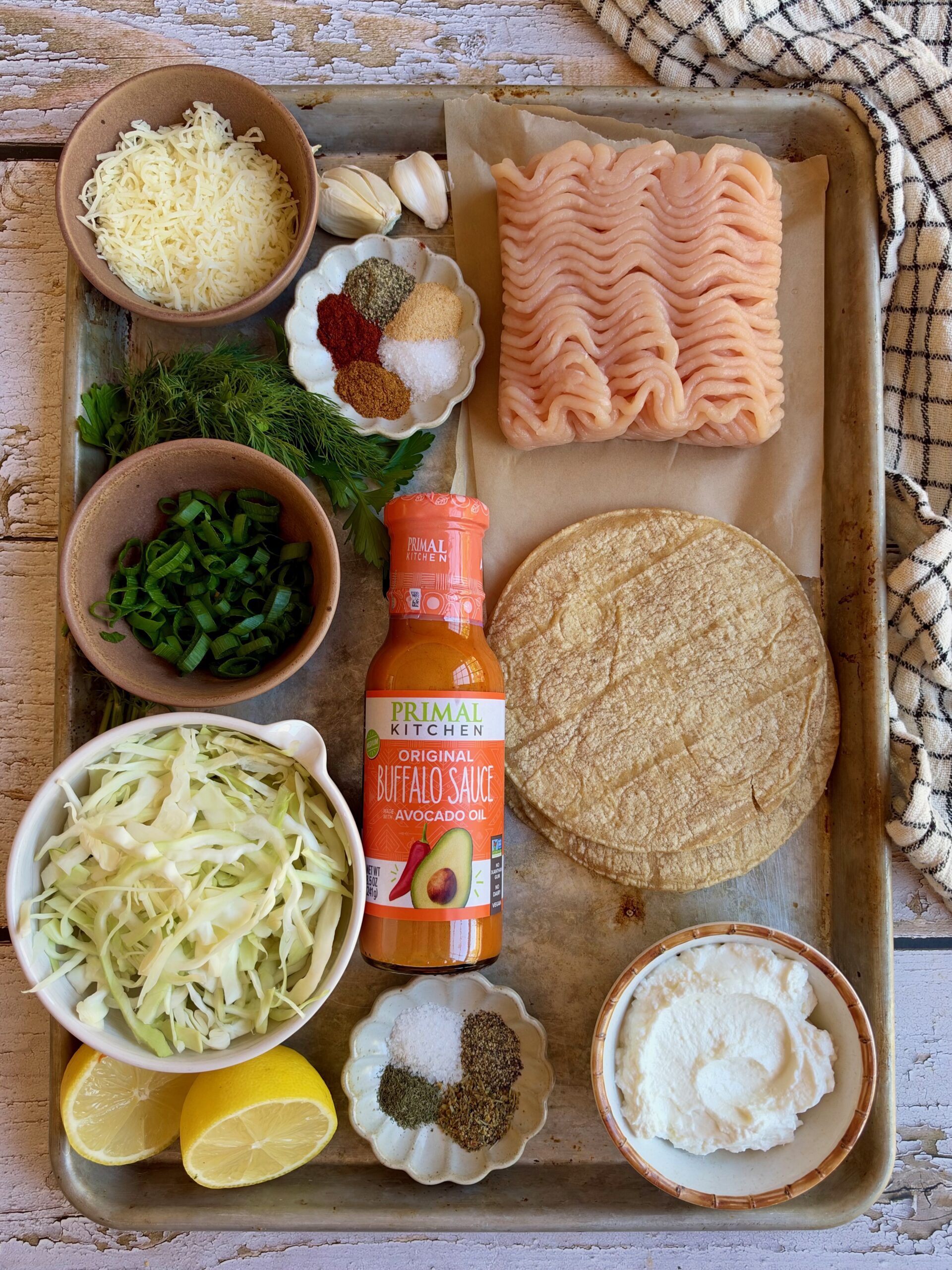 Ingredients for buffalo chicken smash tacos on a sheet pan, including ground chicken, buffalo sauce, corn tortillas, cabbage, Greek yogurt, dill, mozzarella, and spices.