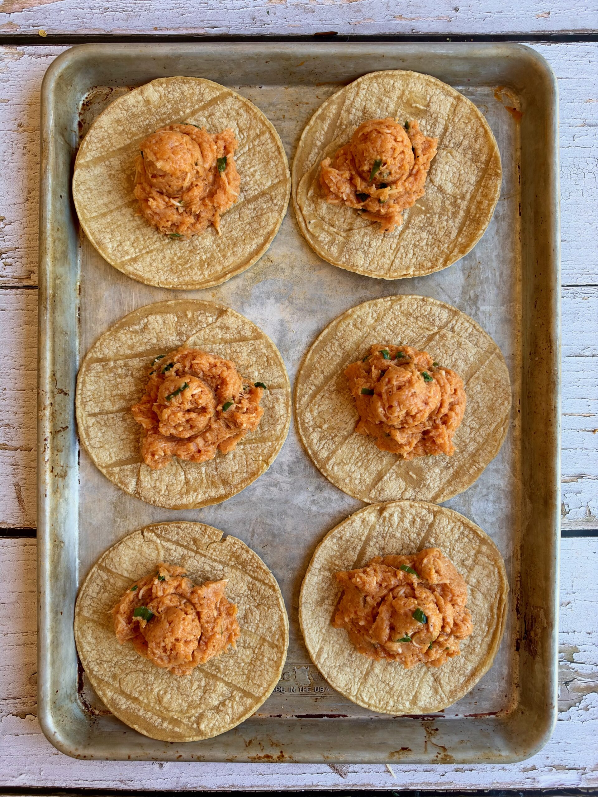 Scoops of buffalo chicken mixture on corn tortillas on a sheet pan.