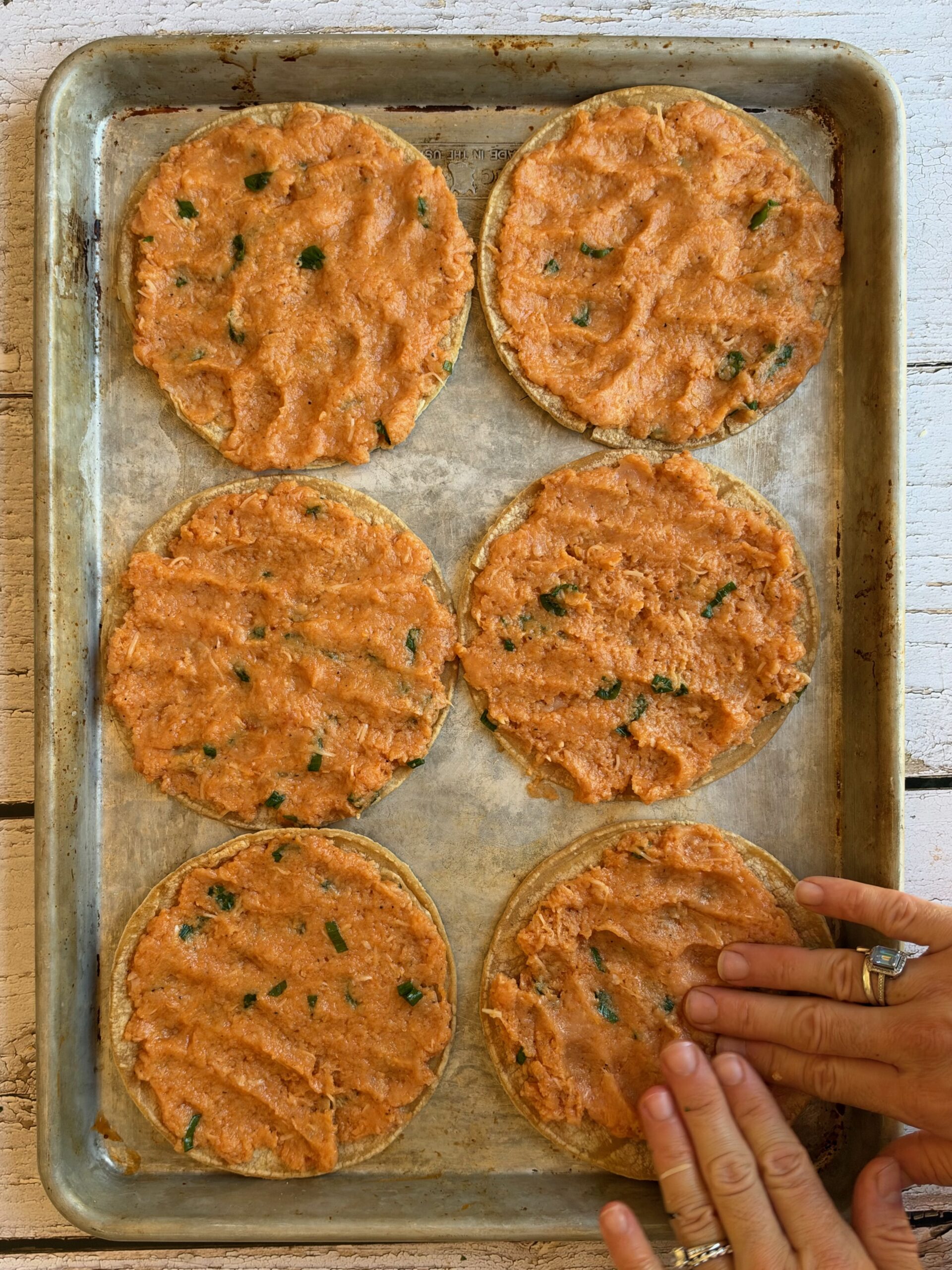 Hands spreading buffalo chicken mixture to the edges of corn tortillas on a sheet pan.