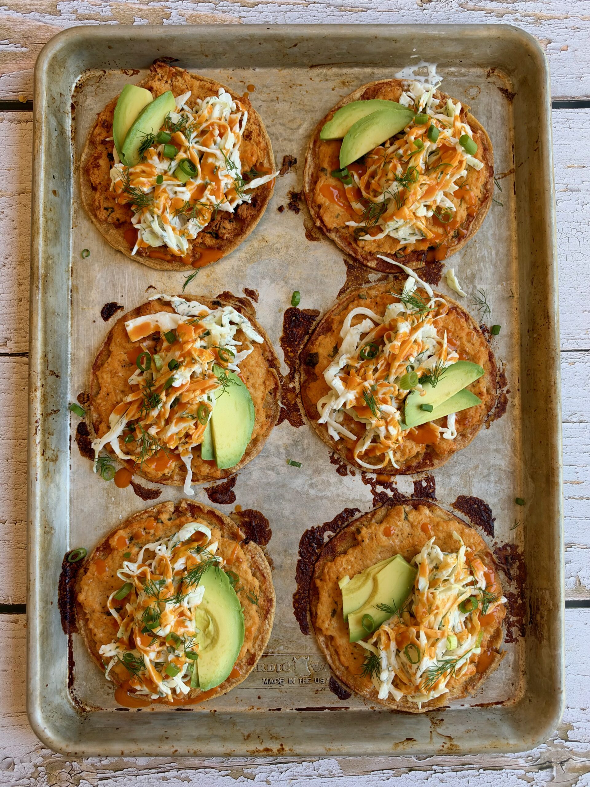 Buffalo chicken smash tacos topped with dill slaw, avocado, and buffalo sauce on a sheet pan.