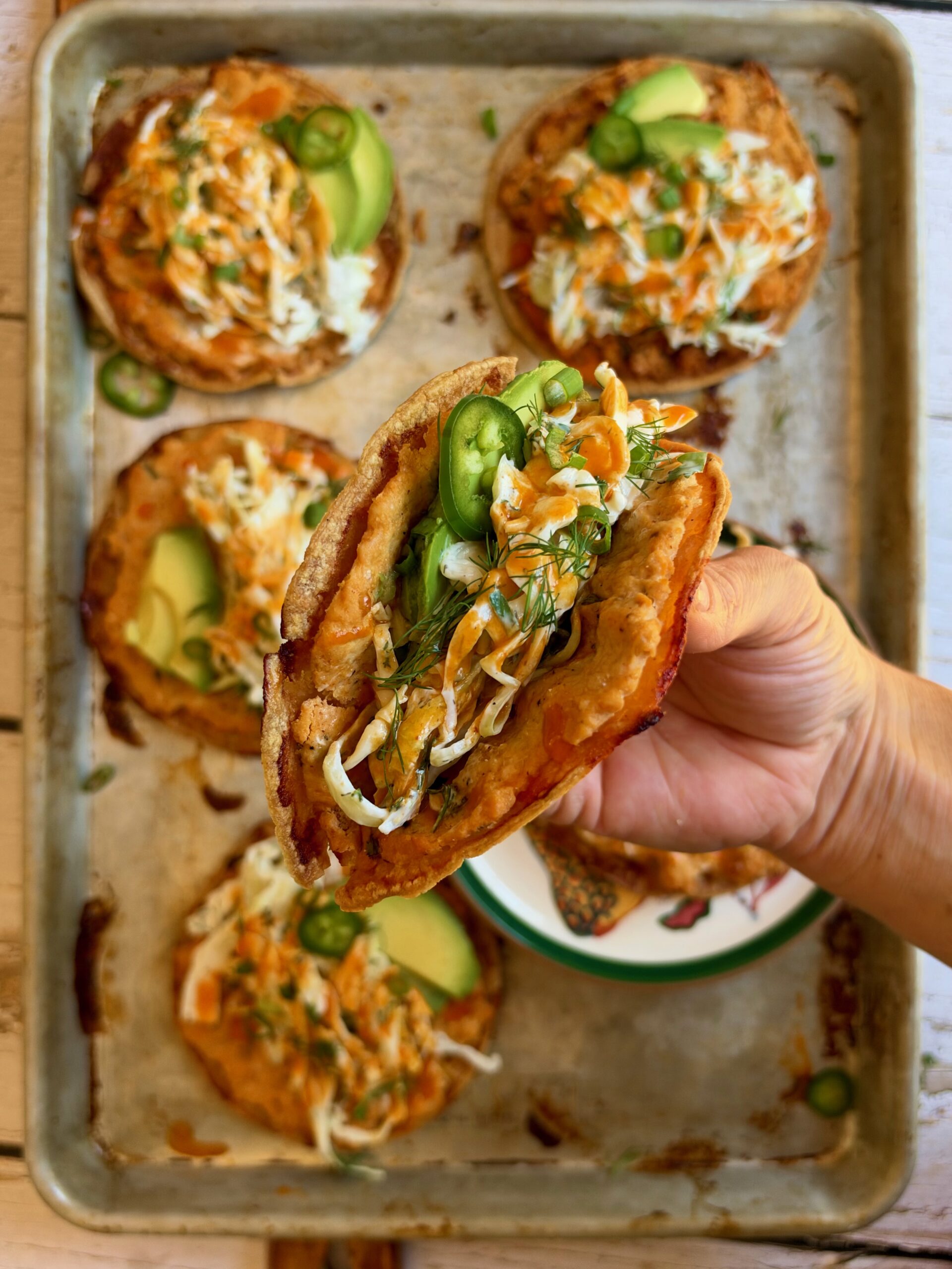 A hand holding a folded buffalo chicken smash taco topped with dill slaw, avocado, and jalapeño.