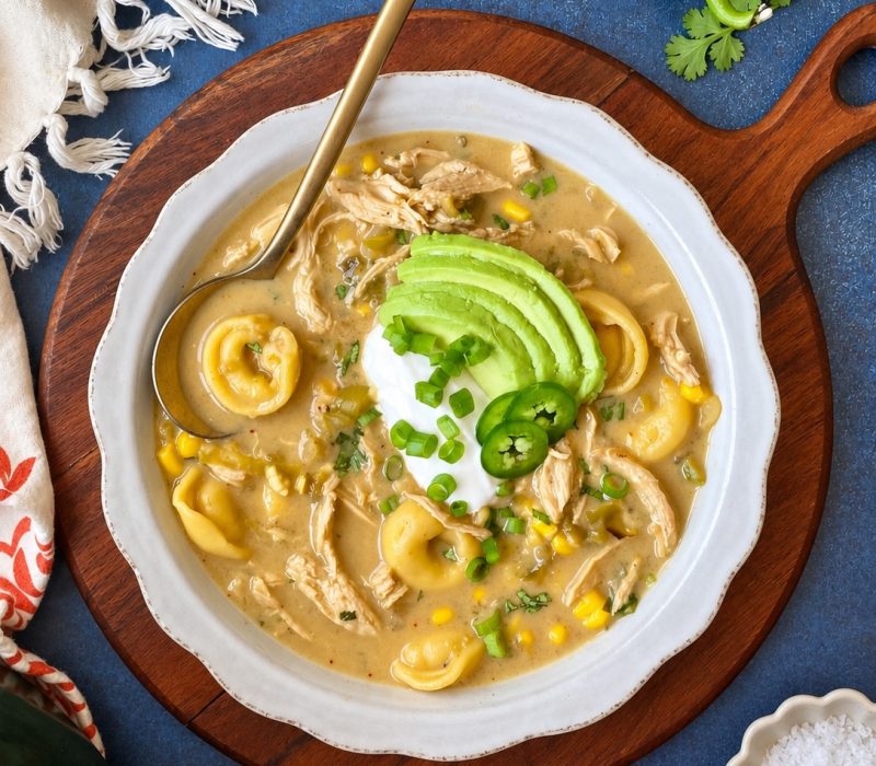 A bowl of creamy soup with tortellini, chicken, corn, sliced avocado, jalapeño, green onions, and a dollop of sour cream, served on a wooden board with limes and peppers nearby.