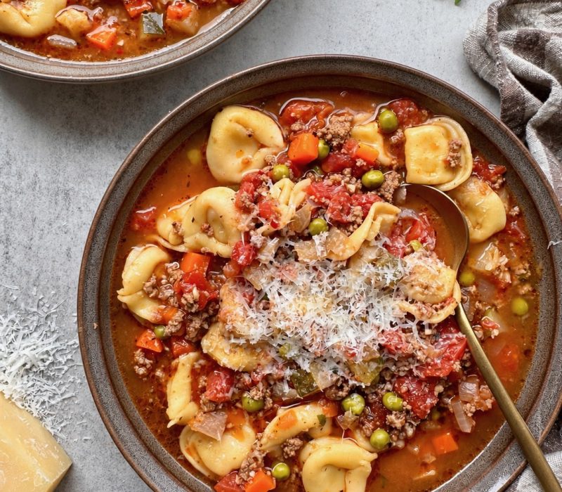 Beef and tortellini vegetable soup topped with grated Parmesan in a dark bowl.