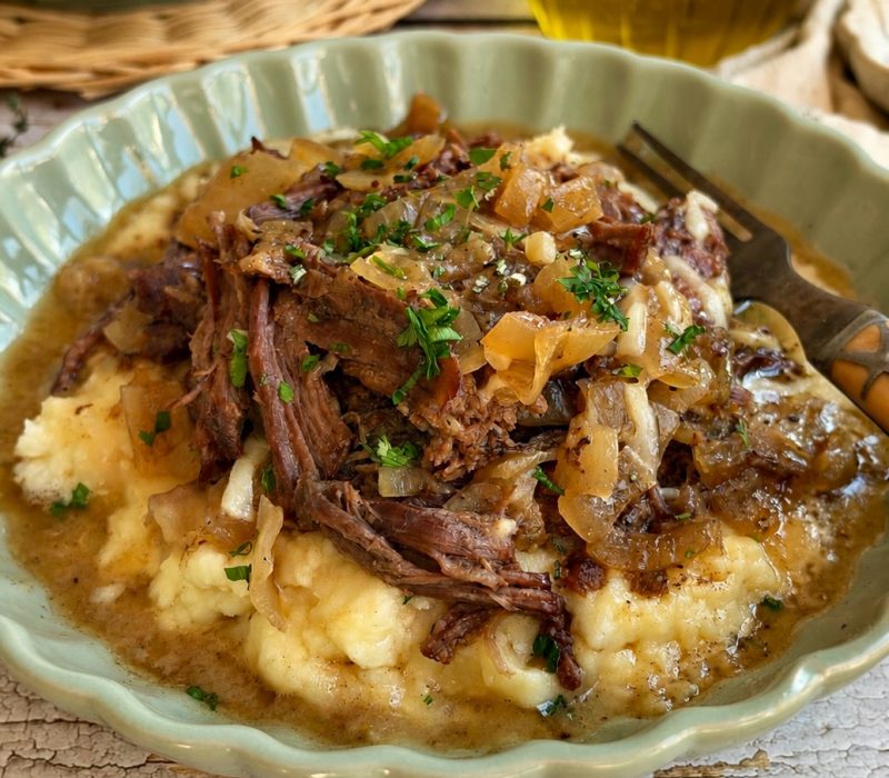Crockpot French onion pot roast with shredded beef and caramelized onions served over mashed potatoes.