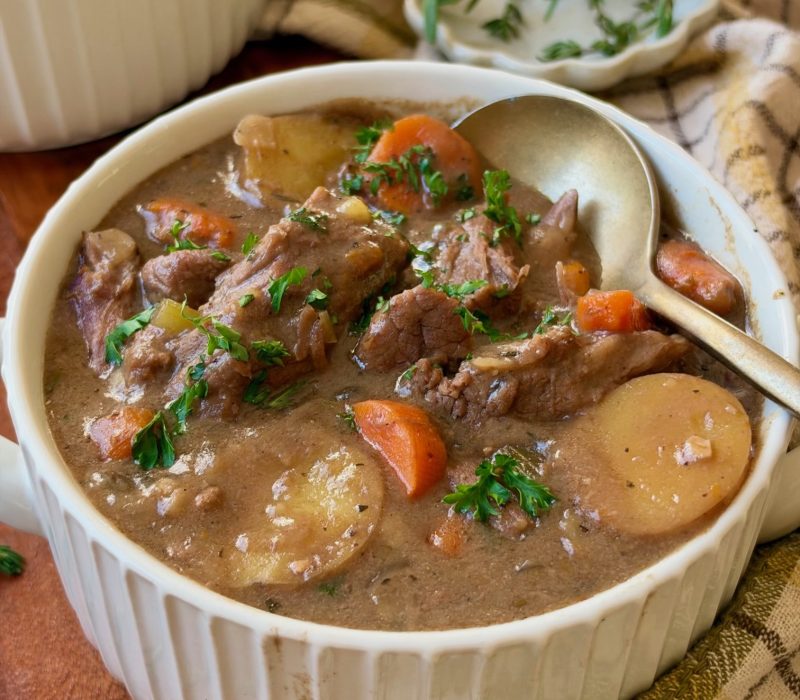 A bowl of hearty beef stew with chunks of beef, potatoes, carrots, and herbs, served with a spoon and garnished with fresh parsley on a wooden table with a plaid cloth and fresh rosemary nearby.