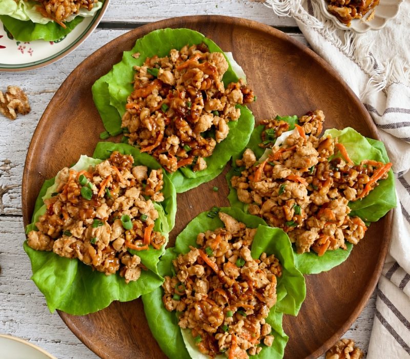 Four lettuce wraps filled with ground chicken and carrots, arranged on a wooden platter.