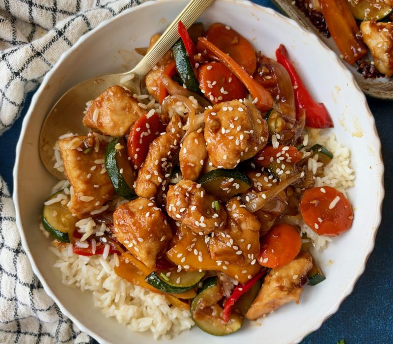 A bowl of chicken and vegetable stir-fry served over white rice, topped with sesame seeds and a gold serving spoon.