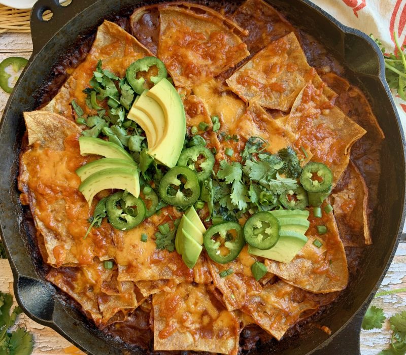 Skillet beef enchiladas topped with melted cheese, avocado, jalapeño, and cilantro in a cast iron skillet.