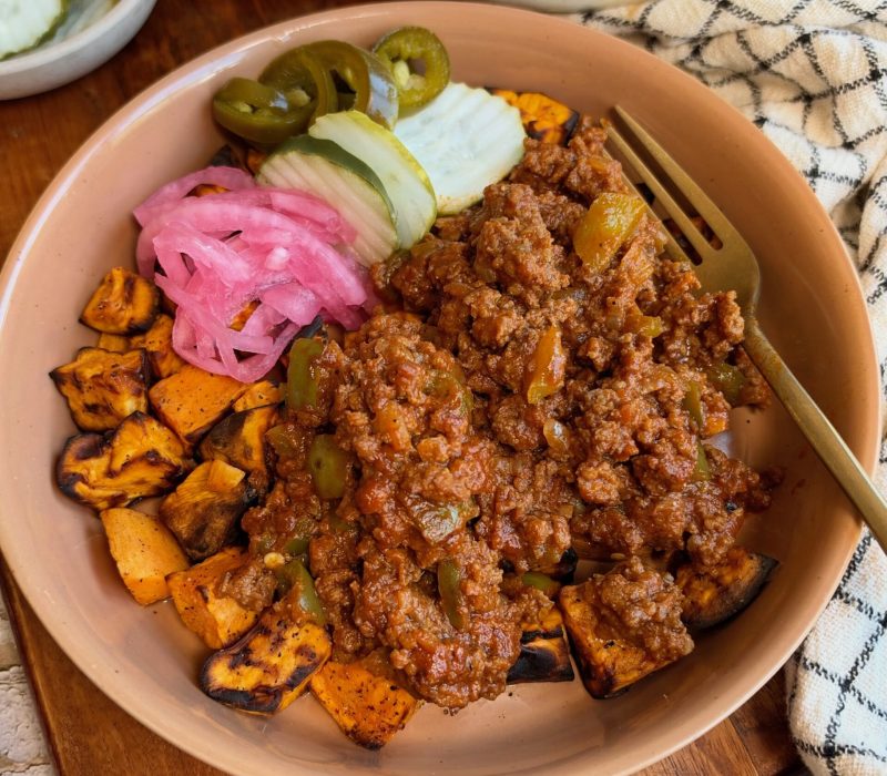 A bowl of ground meat sauce over roasted sweet potato cubes, topped with pickled red onions, sliced cucumbers, and jalapeños, with a fork resting on the side.