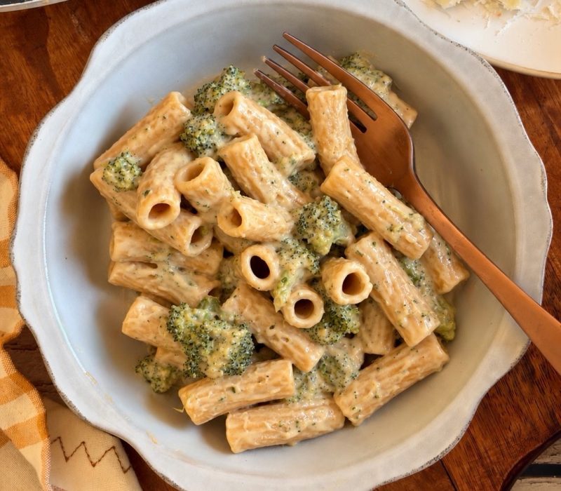 Rigatoni pasta with broccoli in a creamy sauce in a bowl with a fork, next to a block of parmesan cheese.