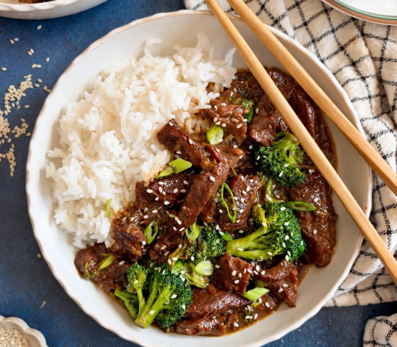 Crockpot Mongolian beef and broccoli served over white rice with chopsticks, topped with sesame seeds and green onions.